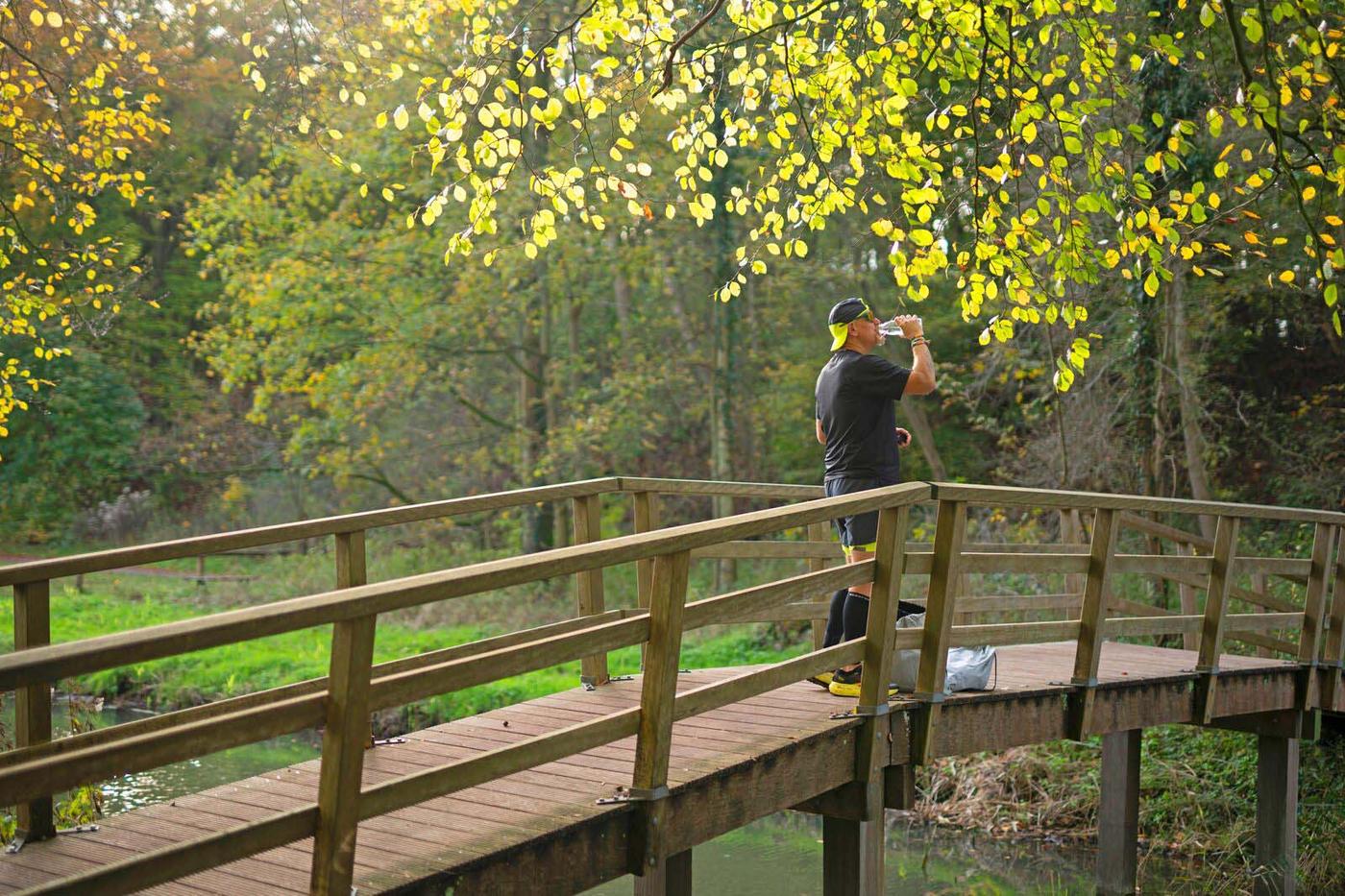 A man stands on a wooden bridge in a forest and drinks water.