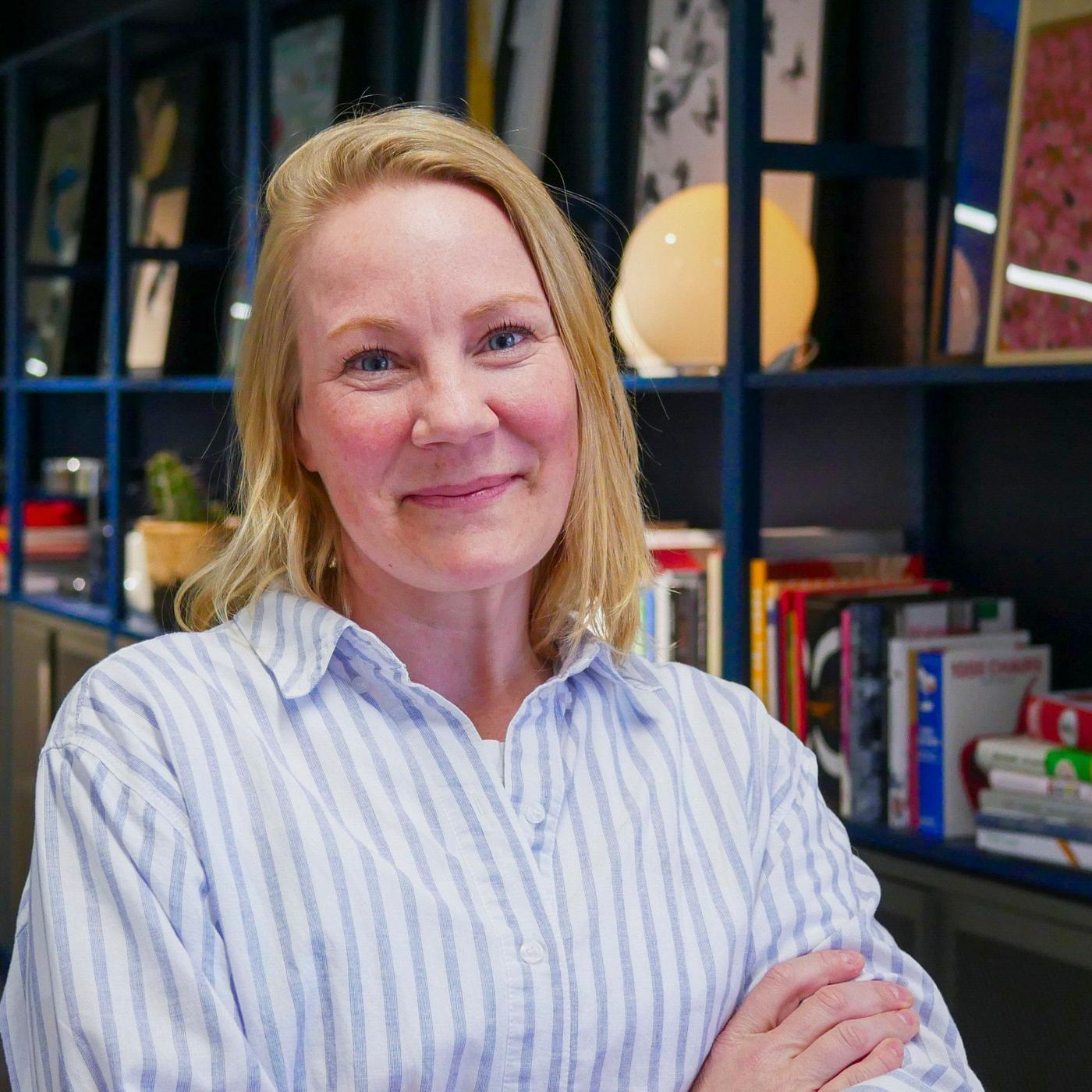 A portrait of a women in a striped shirt, standing in front of a book shelf