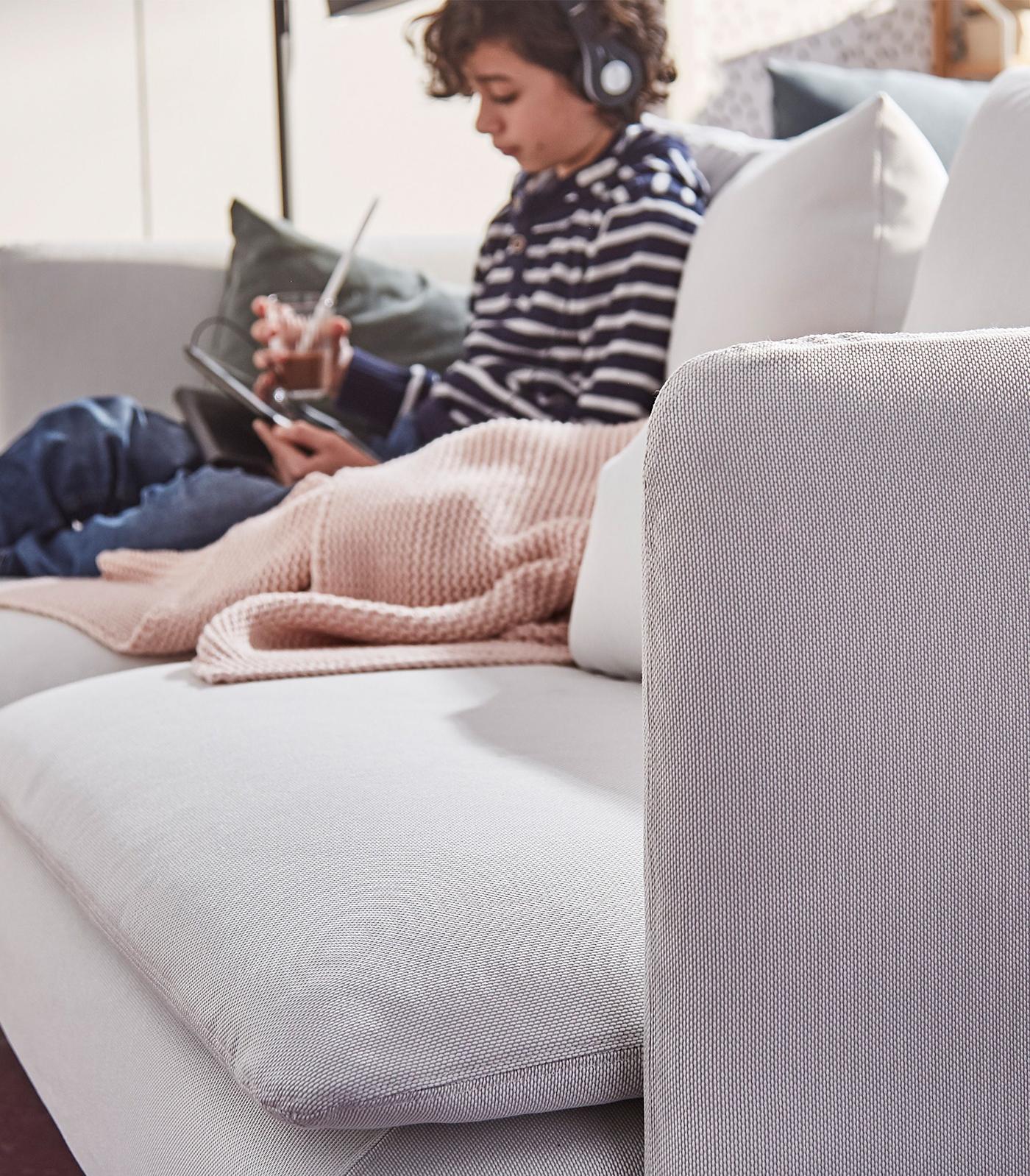 A little boy with headphones sitting on a sofa reading on a tablet.