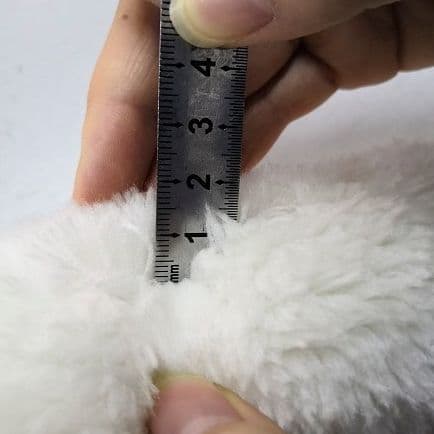 A pair of hands measuring the thickness of a white fake sheepskin with a metal ruler.