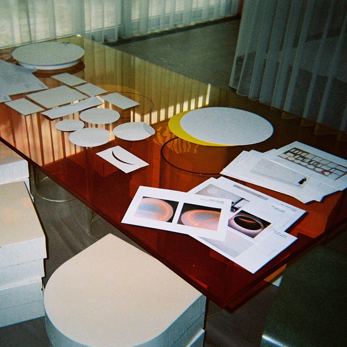 White boxes in piles beside an orange glass table, with a white wall lamp and pieces of paper on top.