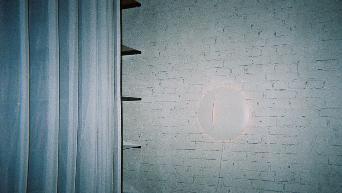 A white round wall lamp on a white brick wall, beside black shelving and a white curtain.