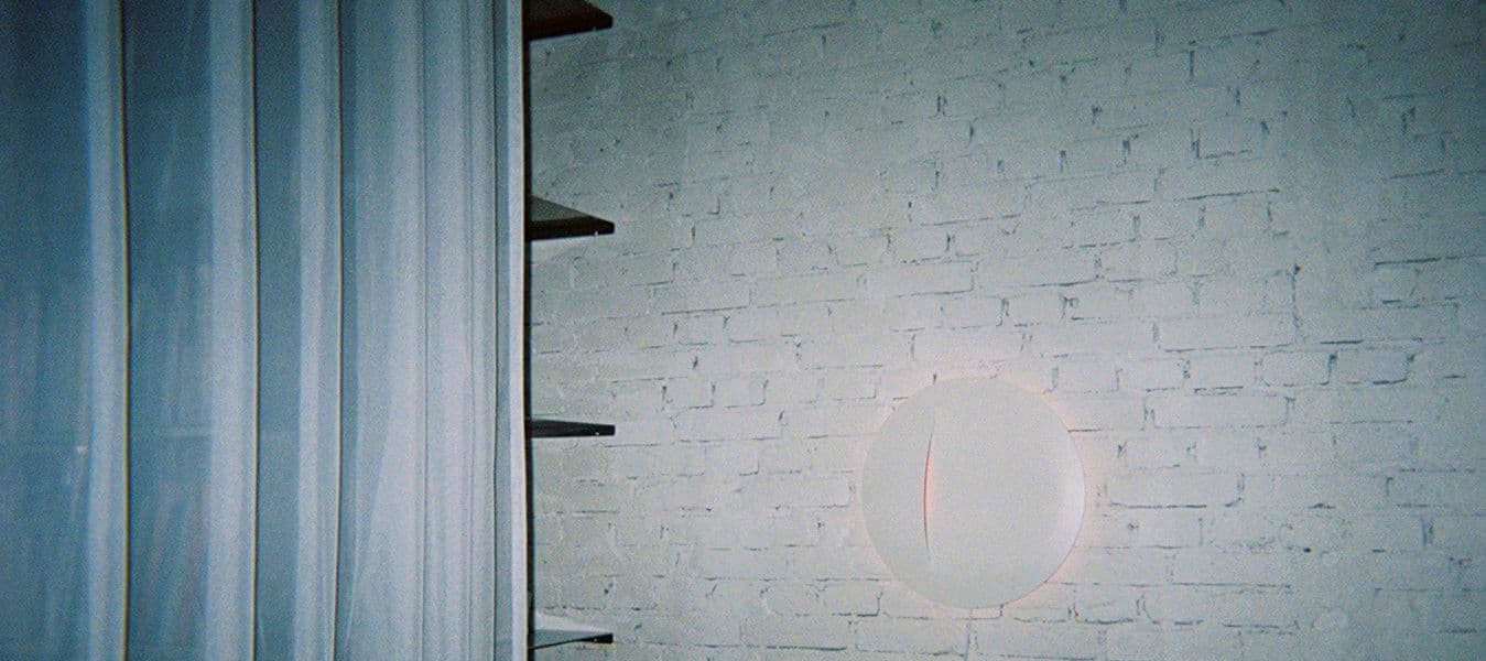 A white round wall lamp on a white brick wall, beside black shelving and a white curtain.