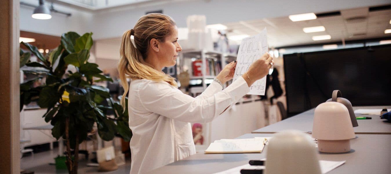 A woman in a white shirt standing at a desk looking at a sketch she's holding in her hand.