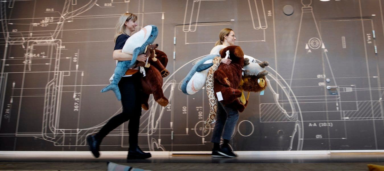 Two women carrying soft toys walking in a hallway.