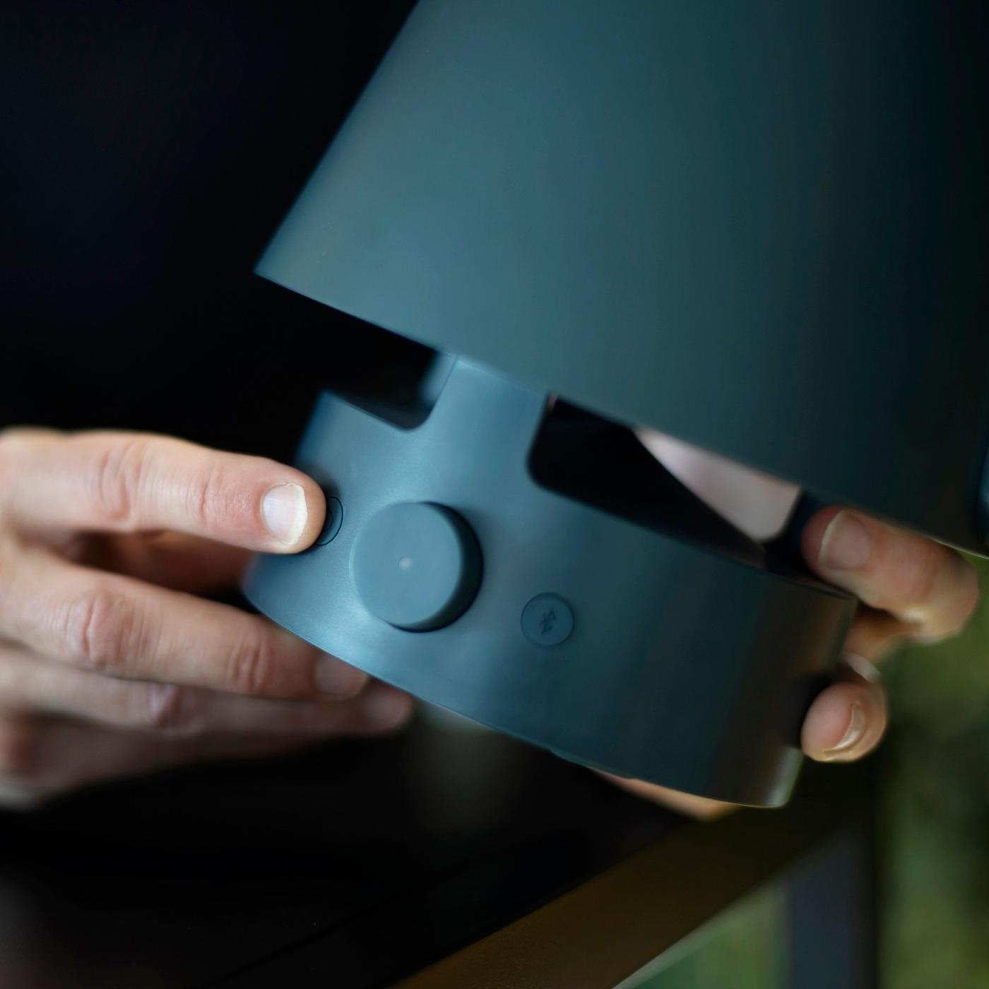 Two hands holding a speaker and a finger pointing at one of the buttons on the speaker