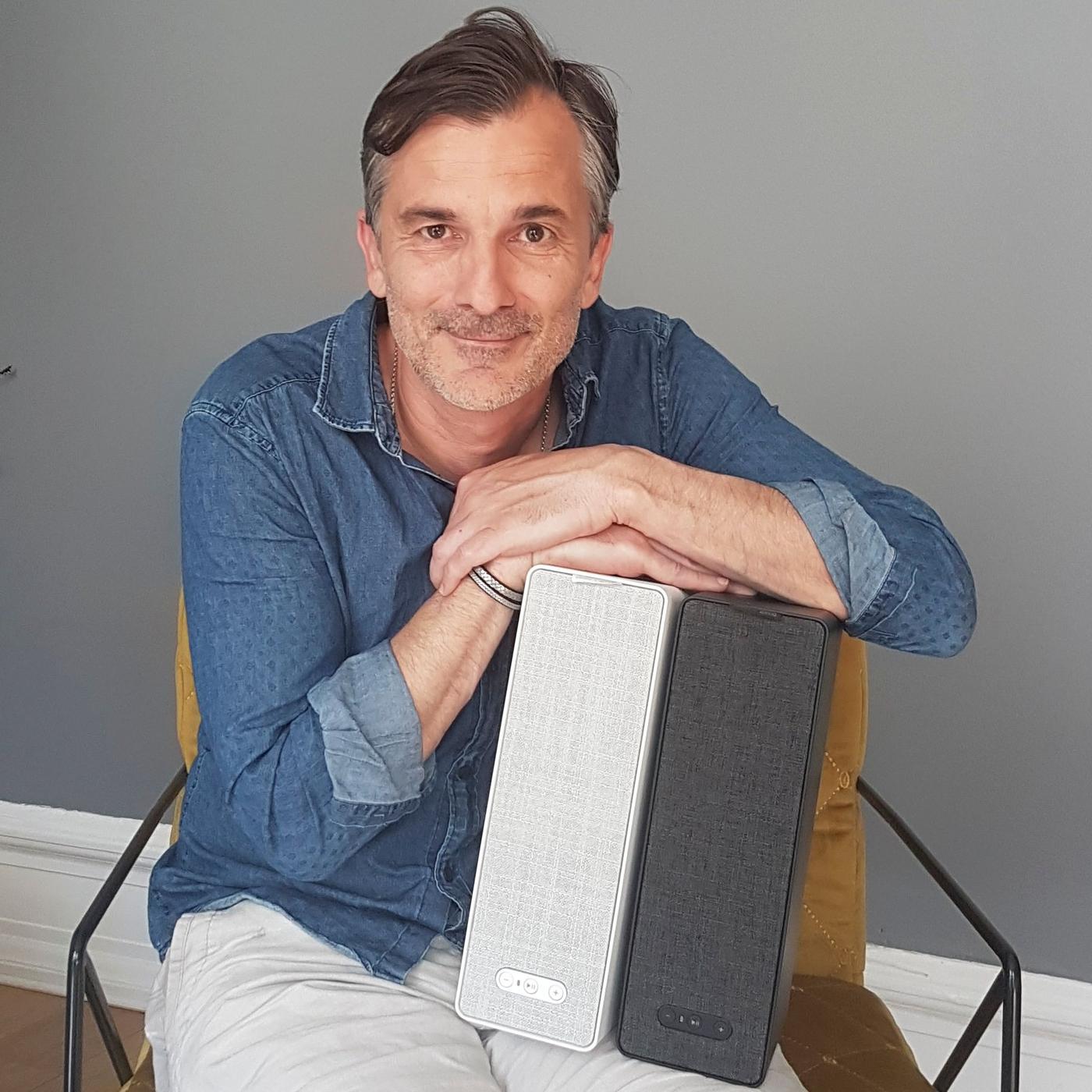 A smiling man with two SYMFONISK WiFi bookshelf speakers on his lap.