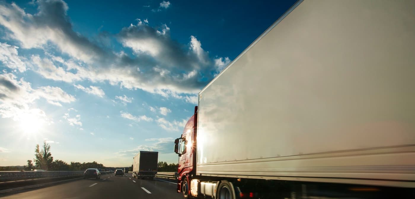 A red cargo truck driving in the right lane on a highway. The sun is setting.