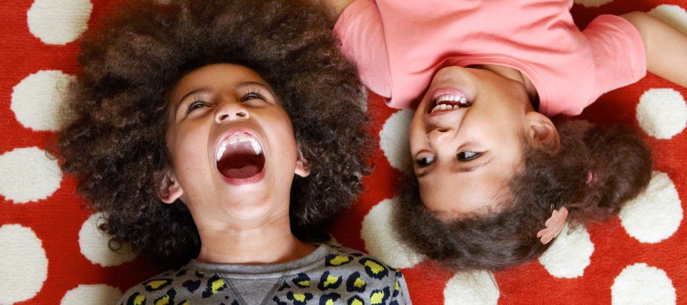 Two girls laughing while lying on a red rug with white dots, their heads close together.