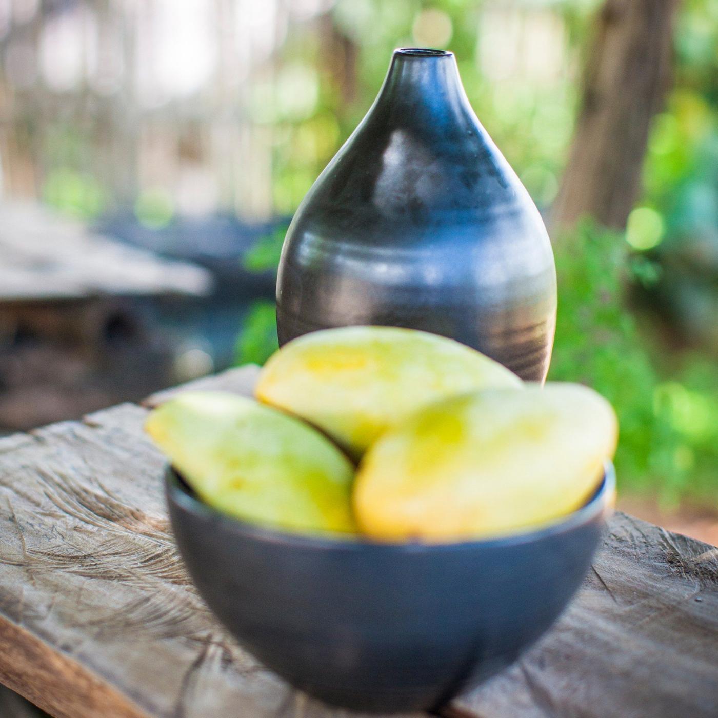 Mango fruit in a handmade, dark brown ceramic bowl and a vase, both from the VERKAN collection, standing on a piece of wood.