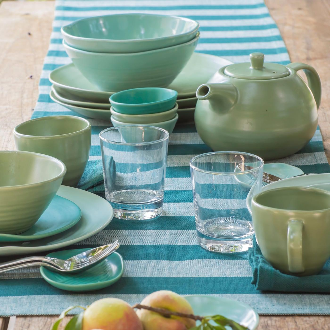 Ceramic VÄLBALANS plates, mugs, bowls and tea pot in different shades of turquoise and green, glasses and cutlery on a table.