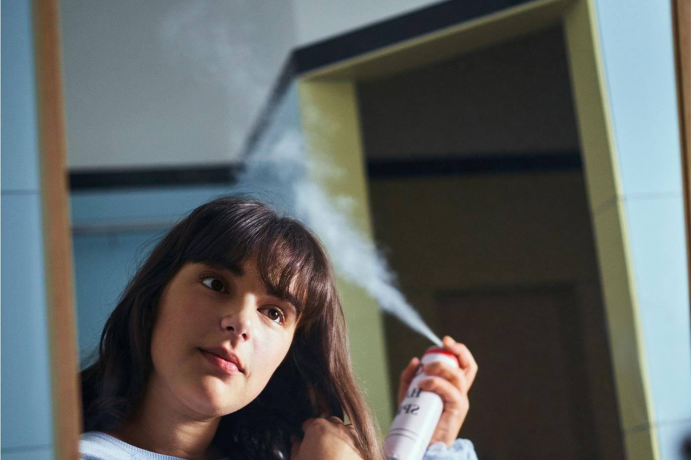 A young woman with dark hair stands in front of a mirror and looks at her reflection while using hairspray.