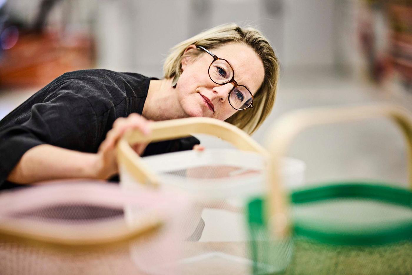 The designer Wiebke Braasch leans down to examine the wooden handle of a white RISATORP basket.