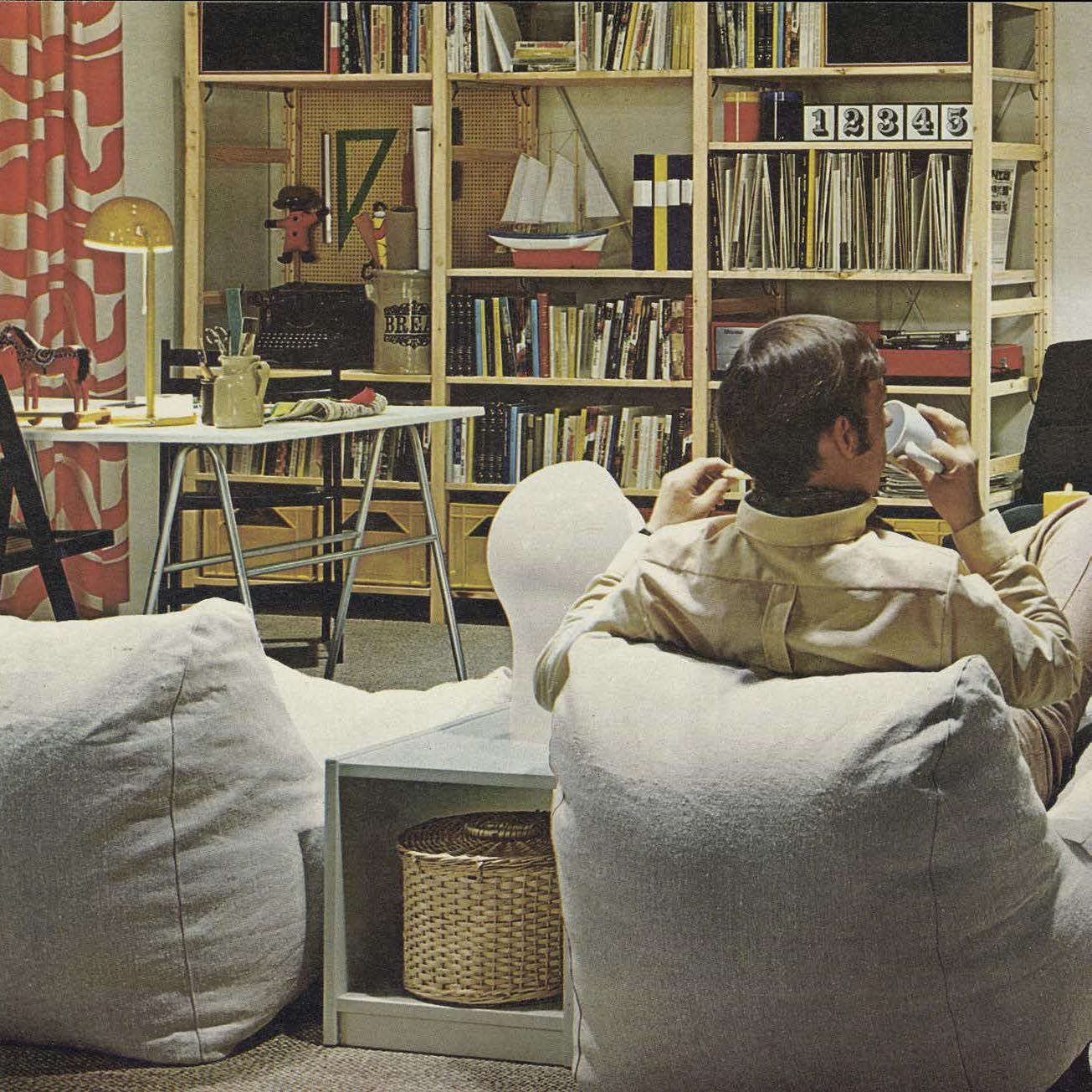 A man sits in a living room in front of a shelving unit filled with books.