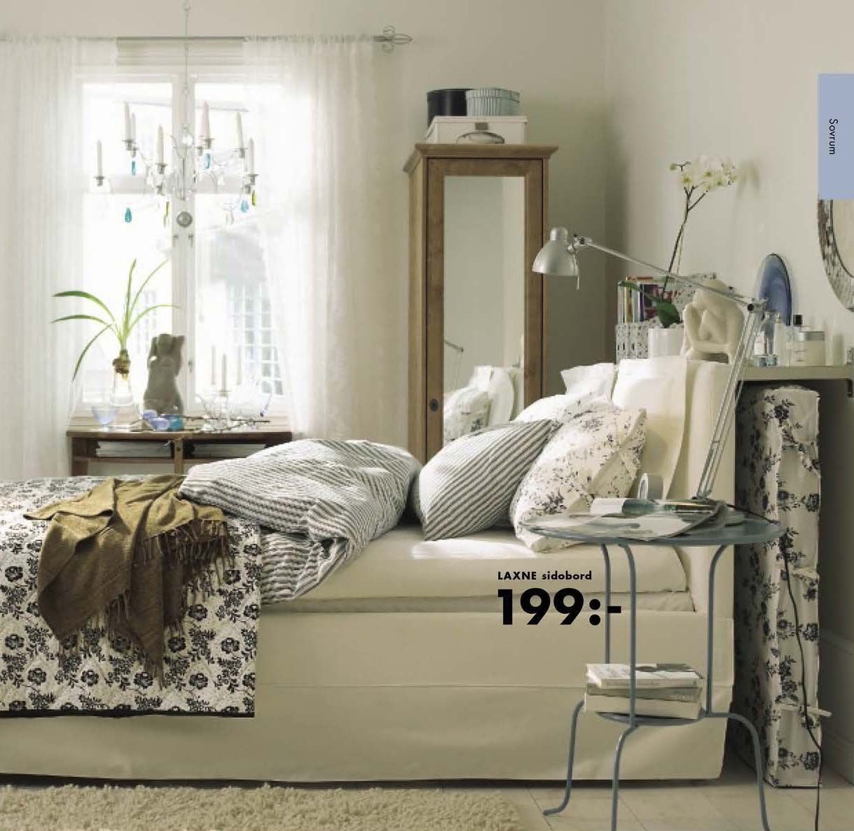 An image from the 2005 IKEA catalogue showing a bed with lots of covers and cushions. In front is a metal table with a lamp and behind is a window and a wardrobe with a mirror.