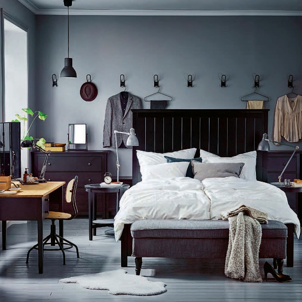 The 2017 IKEA catalogue showing a grey bedroom with a dark grey bed with white and grey bedlinen. Clothes hang from hooks on the wall behind the bed. 