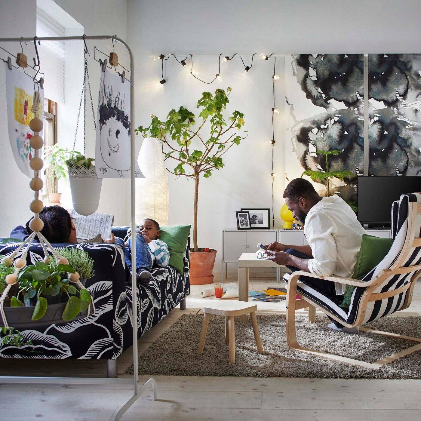 Two adults and a child relax in a creative, art-filled living room with a KLIPPAN sofa and POÄNG armchair.