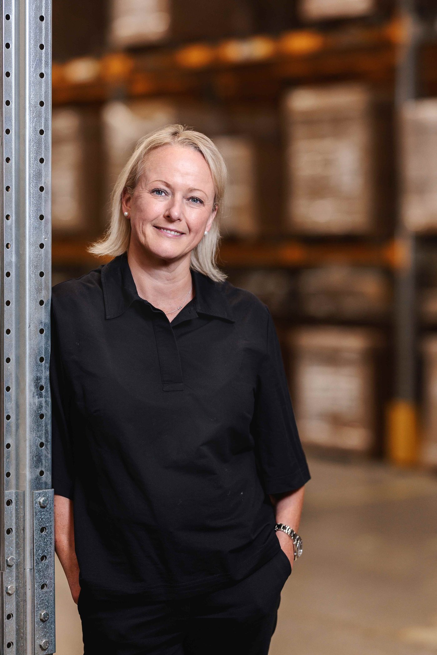 Portrait of Susanne Waidzunas leaning on a metal shelving system in an IKEA store. She is wearing black clothes and smiling at the camera with her hands in her pockets.