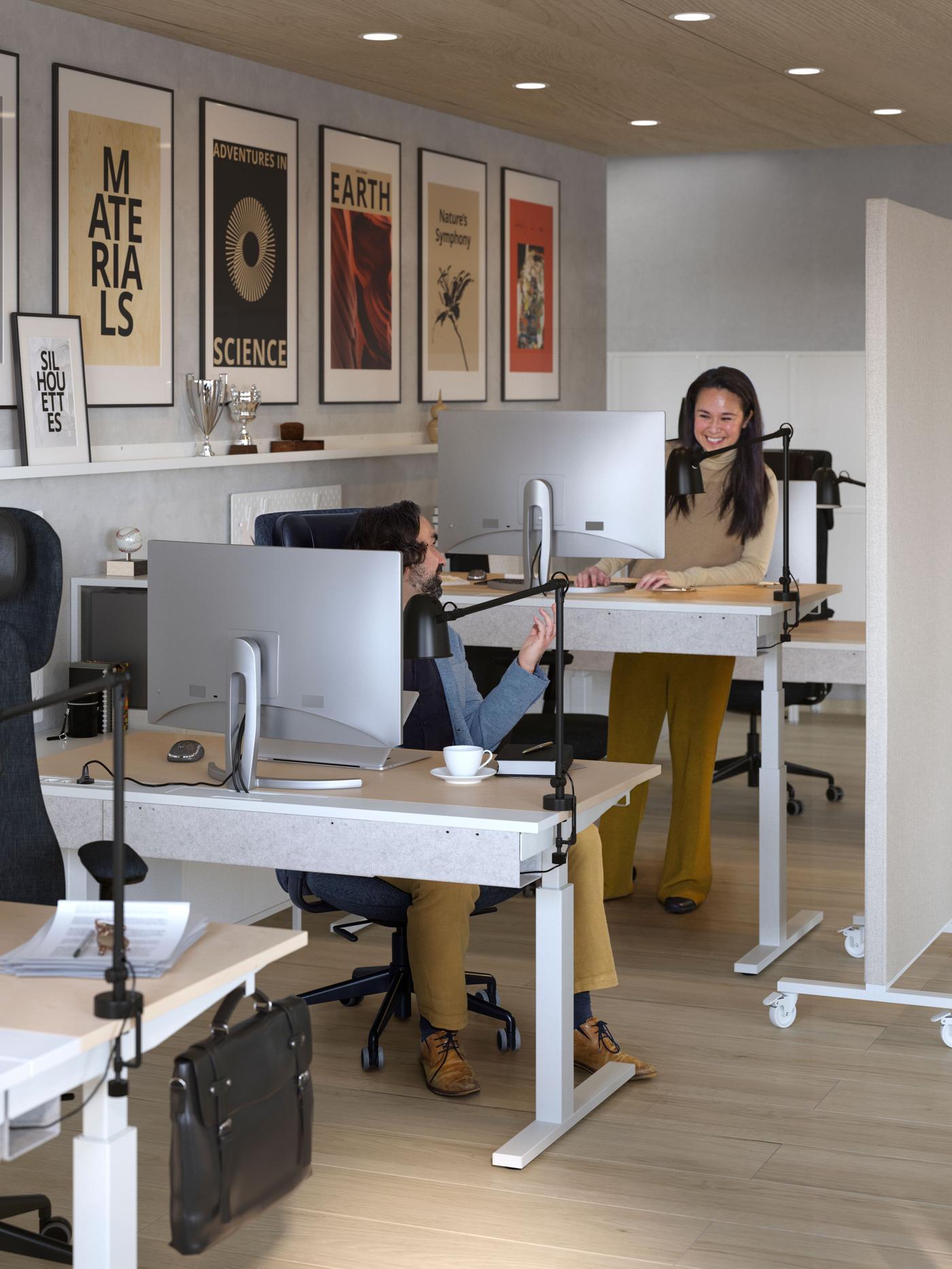 Office space, two people that are both sitting behind a desk with computers. They are looking at each other and laughing. 