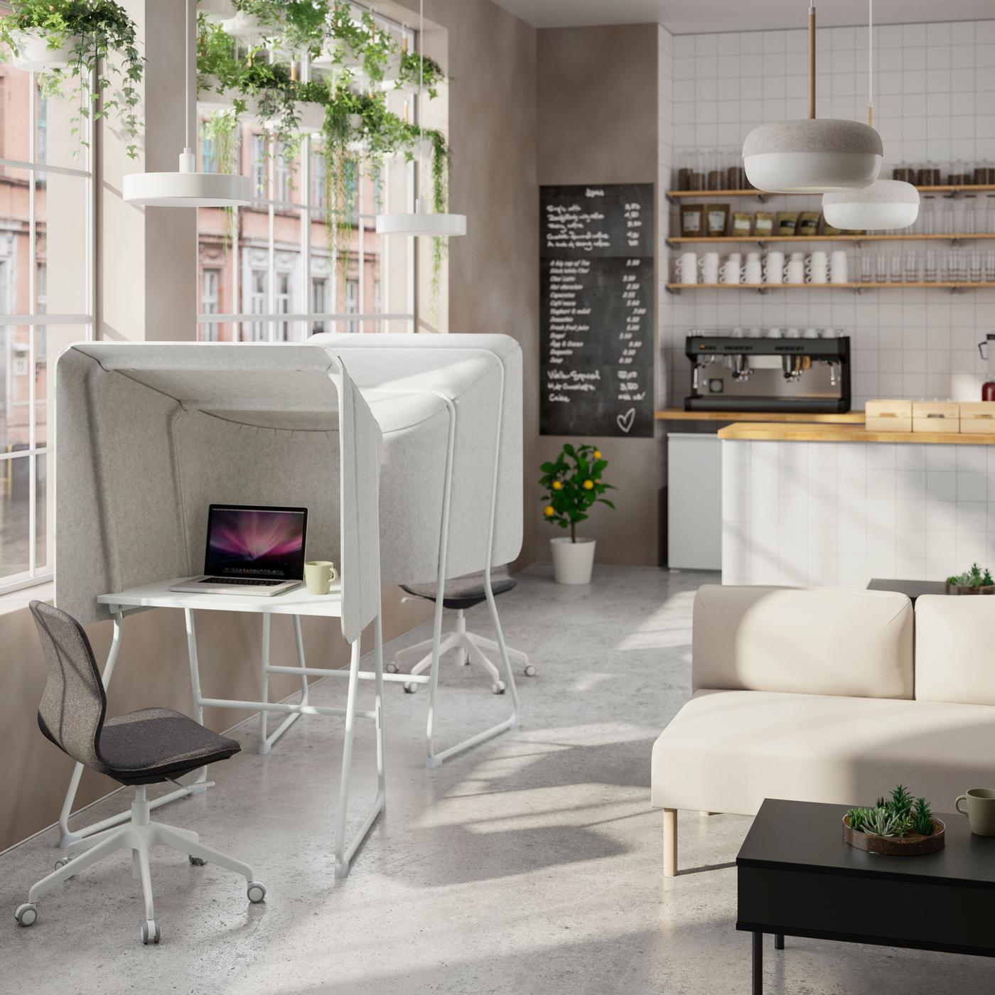 Office coffee area with two cubicle desks, och other. Behind it the bane with a laptop, facing ear, coffee machine and cups, and next to the cubicles a light colored sofa with black table.