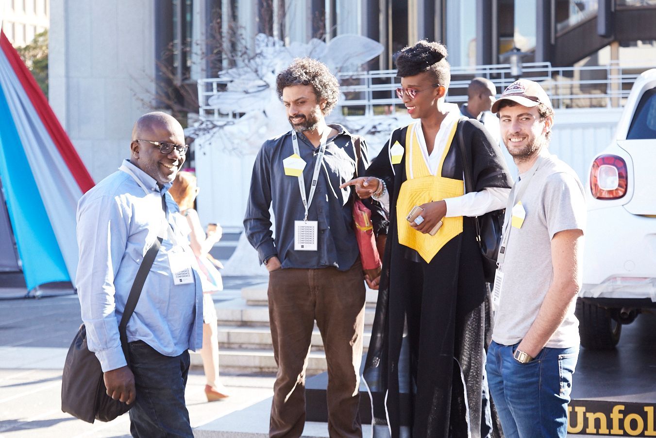 Four people stand outside. One looks at the camera, while the other look at one of the people.