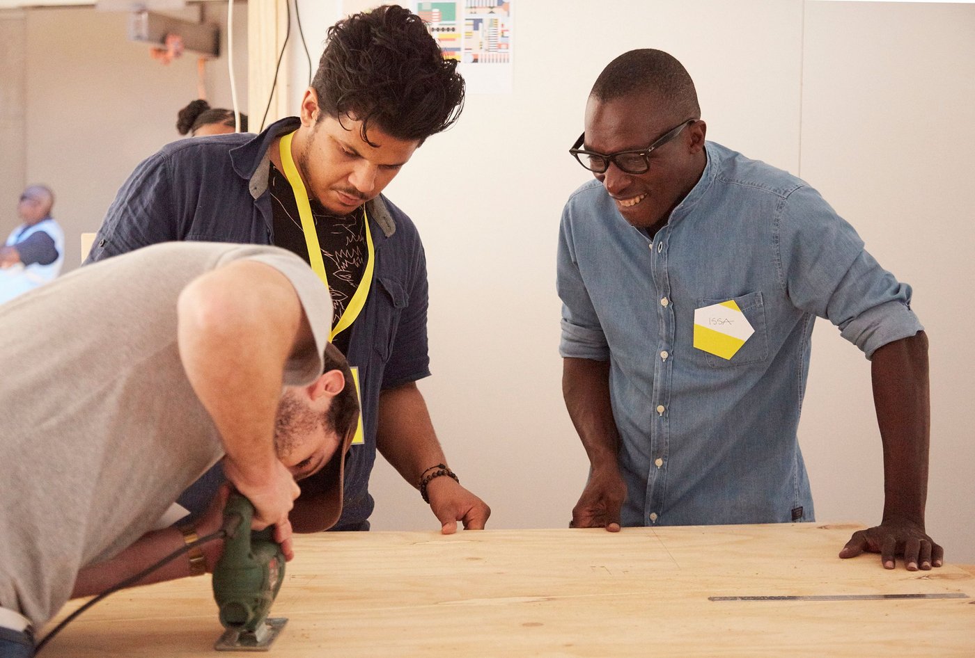Two men watch another man using an electric tool.