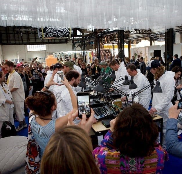 A large group of people surrounding a small group of people in white suits, working at a table.