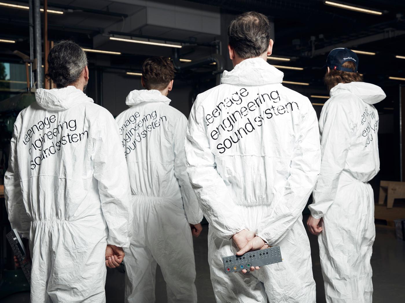 A great of people stand with their backs to the camera. They are wearing white hazard suits.