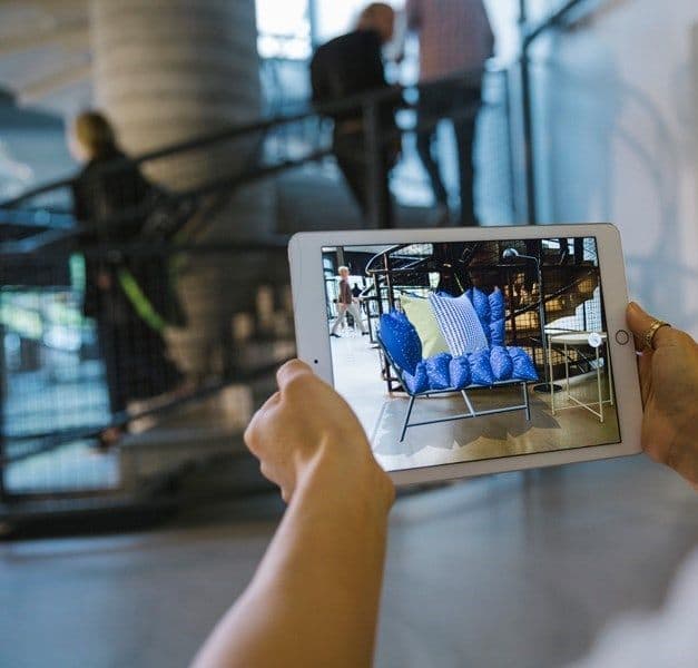 A persons hands holding an iPad which is running an AR furniture-placement app.