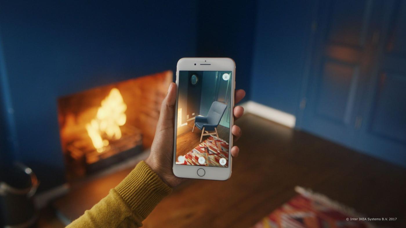 Hands holding a phone in a room near a fireplace, interacting with the IKEA Place app.