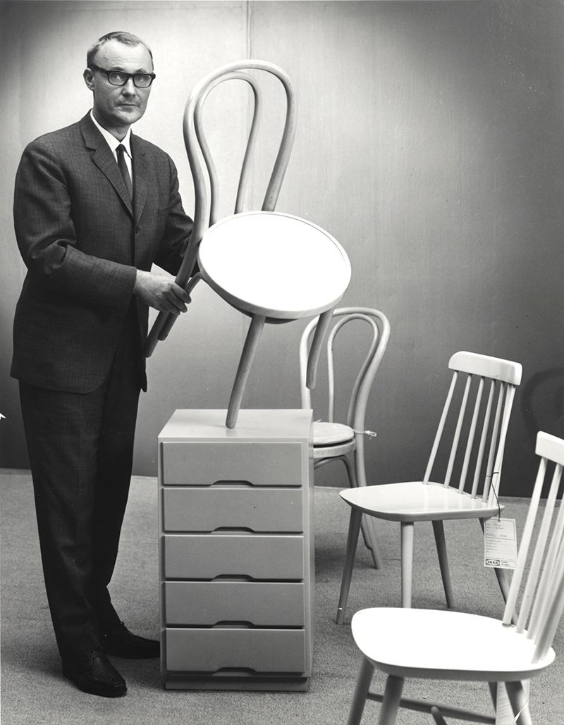Black and white photo of Ingvar Kamprad holding a chair, with chairs and a chest of drawers on the floor around him.