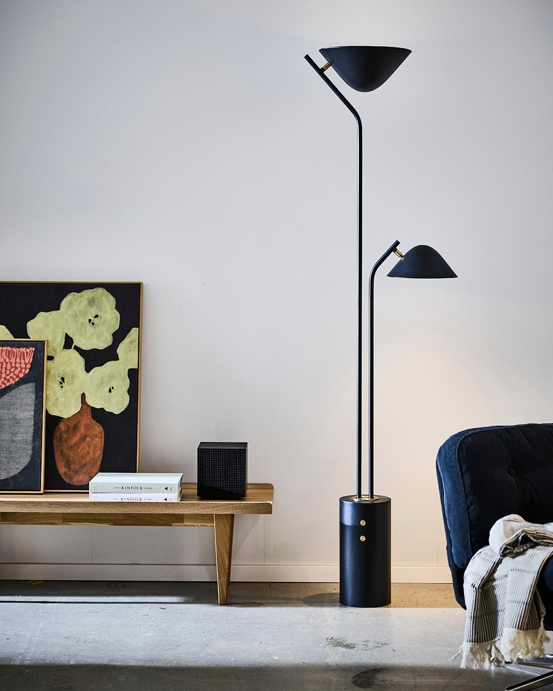 Black floor lamp with dual heads standing in a minimal living space beside a wooden bench, artwork, and chair.