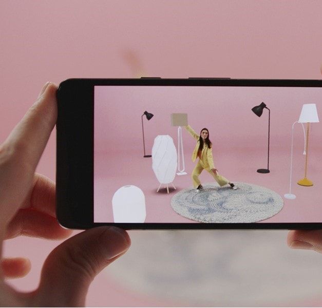Hands holding a smartphone filming a person standing in a pink roomset with a rug and various floor lamps.