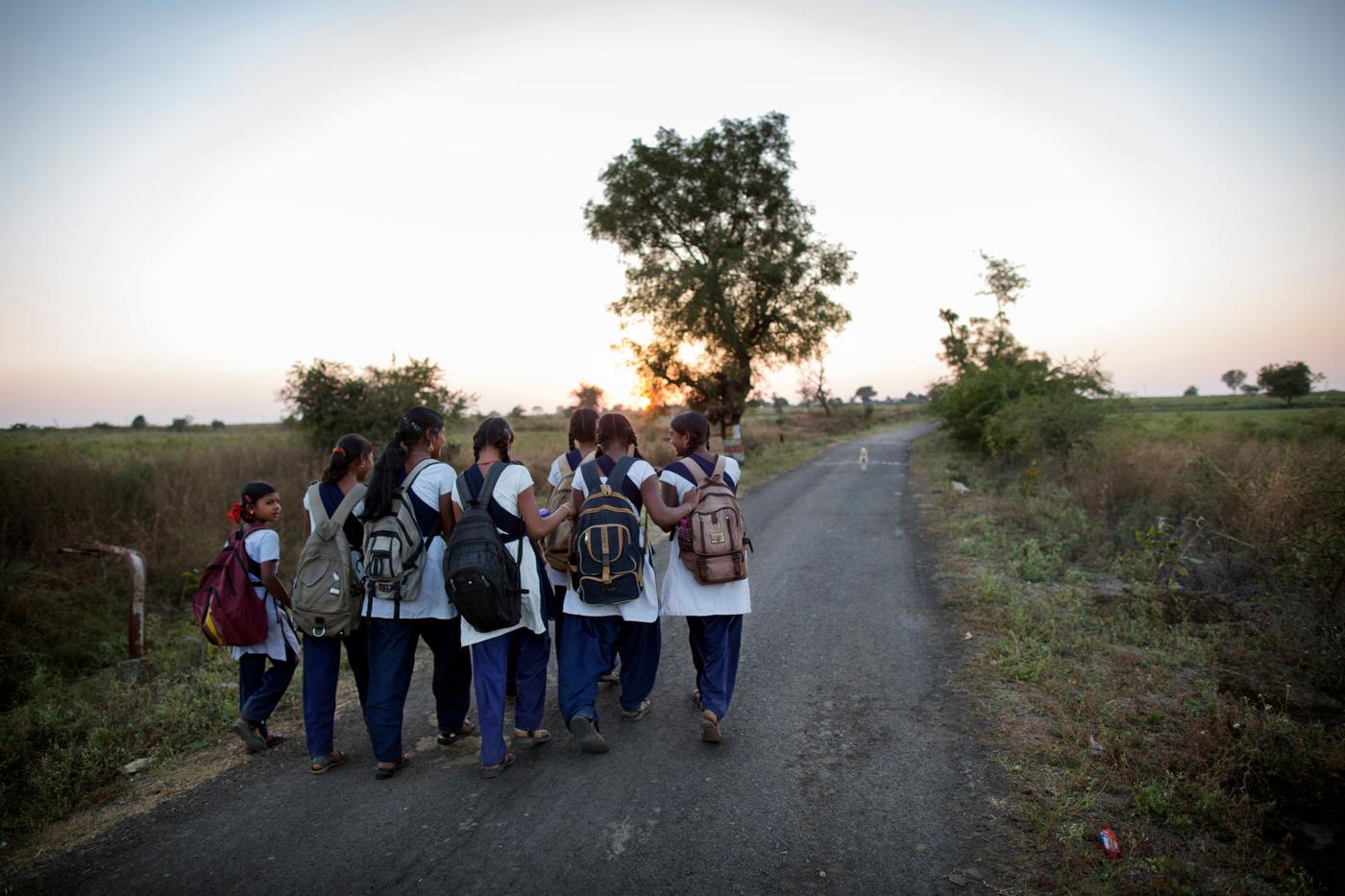A group of children wearing school backpacks, walking along a countryside road in the late afternoon.