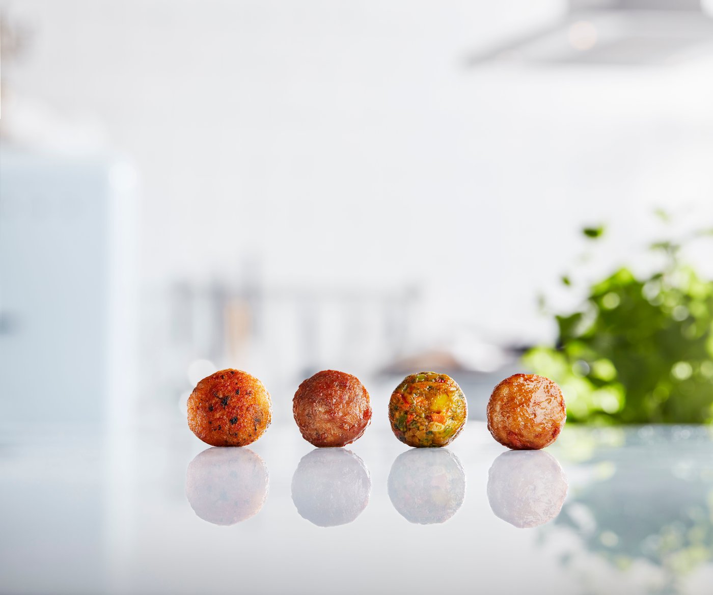 Four different types of IKEA meatballs lined up on a shiny reflective worktop in a white room.