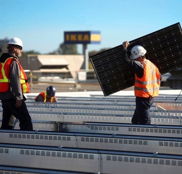 A person carries a solar panel on top of a store roof, while another person watches. A third person is working in the background.