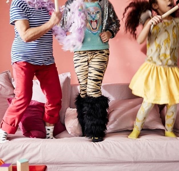 Three children wearing fun, colourful outfits jumping on a pink sofa.