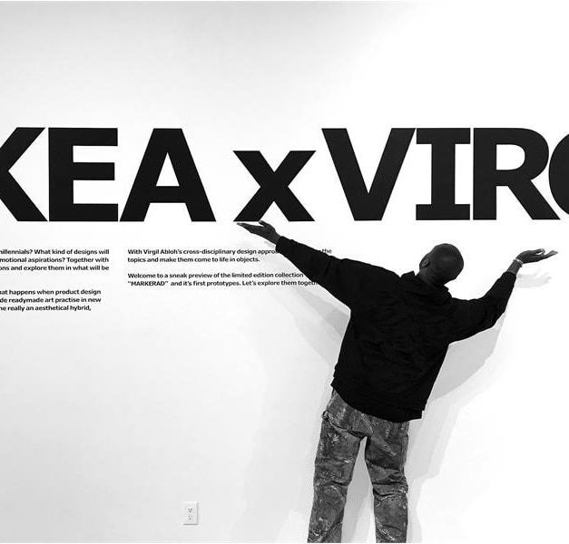 Black and white photo of Virgil Abloh standing with arms outstretched in front a wall reading "IKEA x VIRGIL"