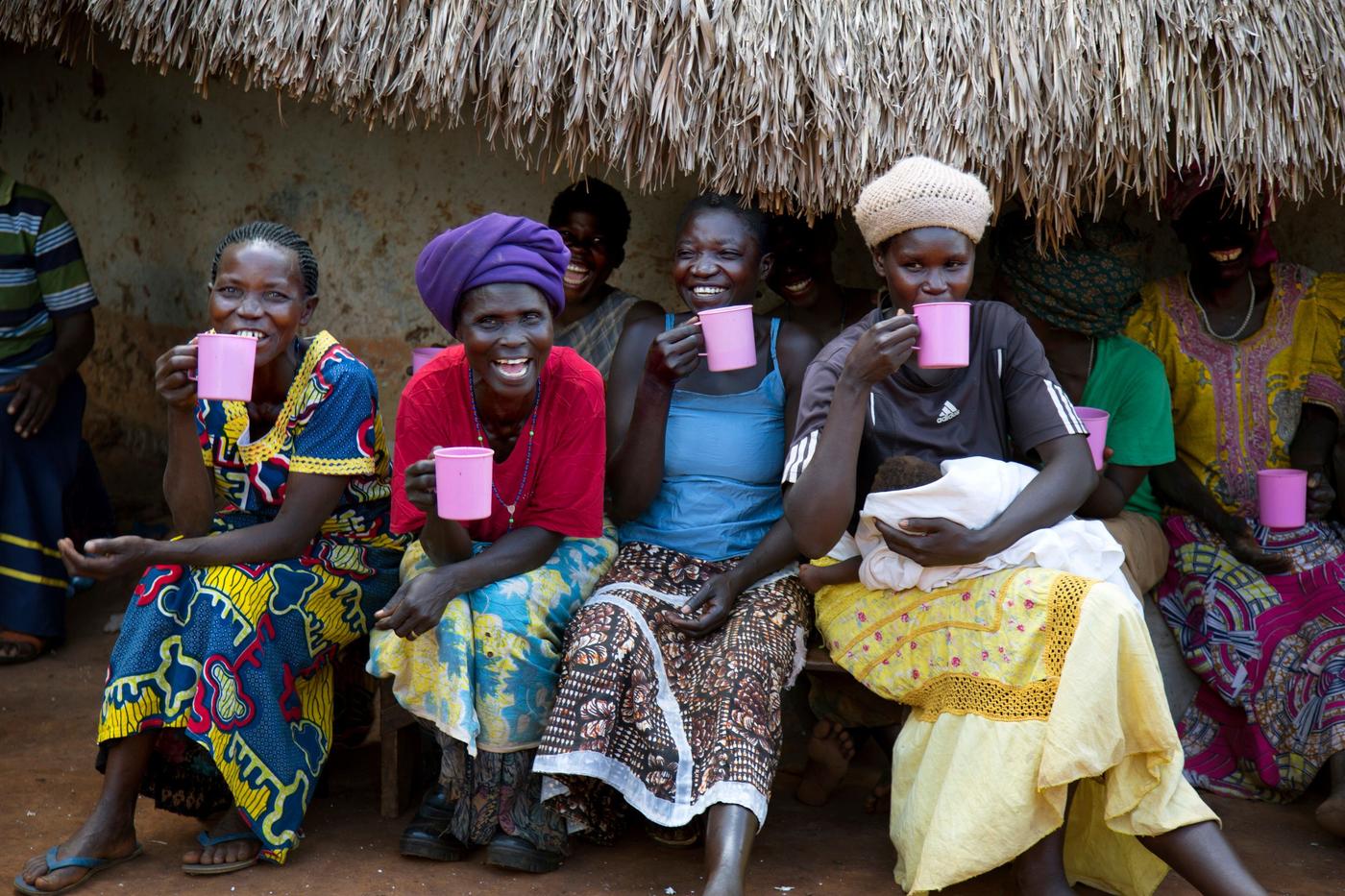 A group of women sitting outside a thatch-roof building and drinking from pink cups.