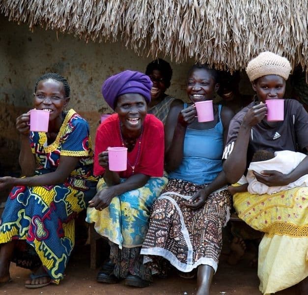 Four people sitting outside a grass-roof building drinking from bright pink cups and smiling.