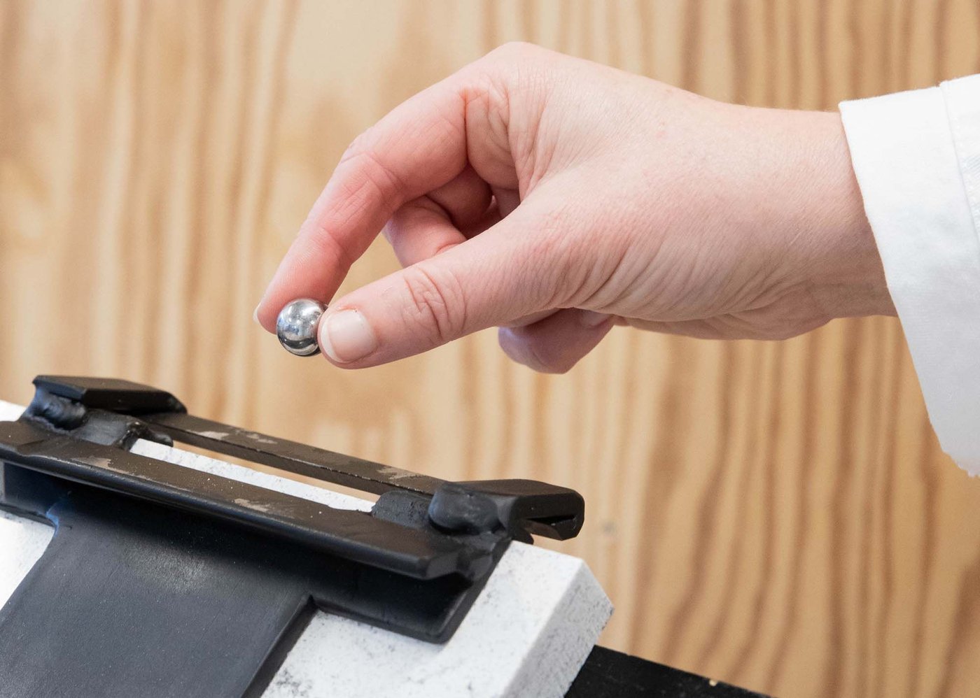 A hand holds a small metal ball above the device used for edge impact testing.
