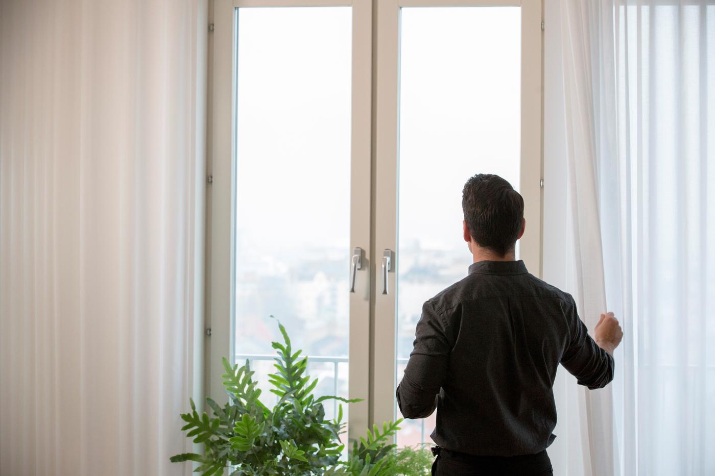 A person stands at the window, next to plants. They are pulling one of the curtains back.