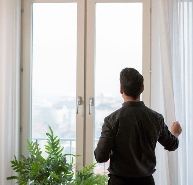 A person stands at the window, next to plants. They are pulling one of the curtains back.