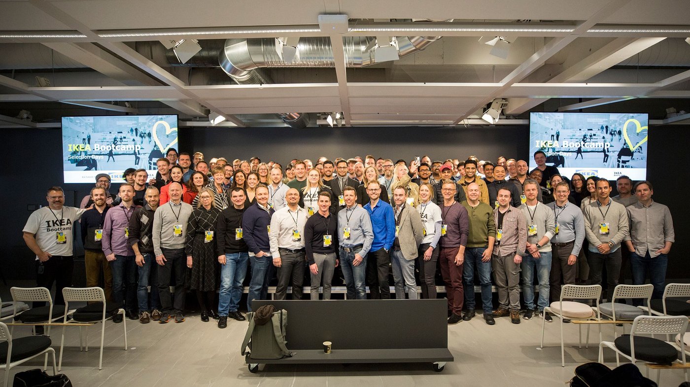 A large group of people stand in rows, looking at the camera. There is a TV screen behind them.