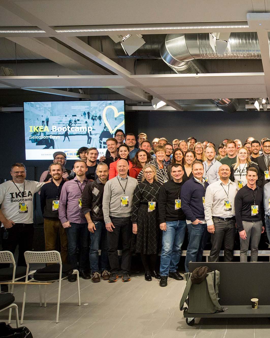 A large group of people stand in rows, looking at the camera. There is a TV screen behind them.