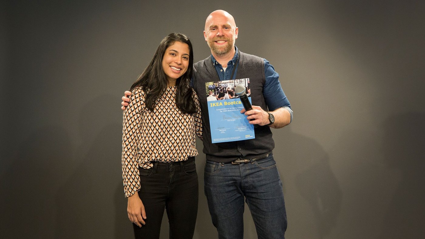 A man and woman smile at the camera. The man is holding a microphone and a certificate.