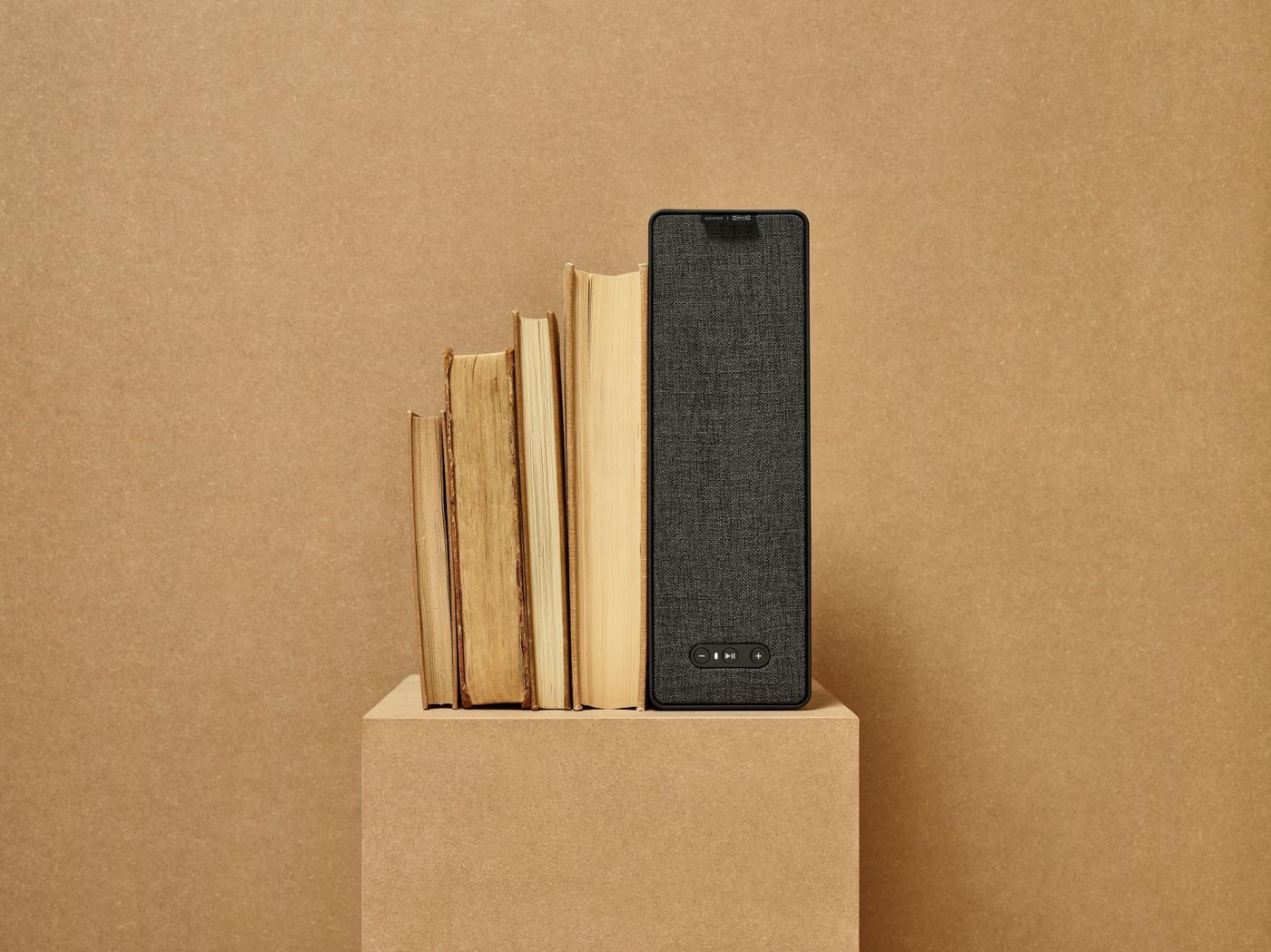 Black SYMFONISK WiFi bookshelf speaker displayed with books on a sandy-coloured plinth and background.