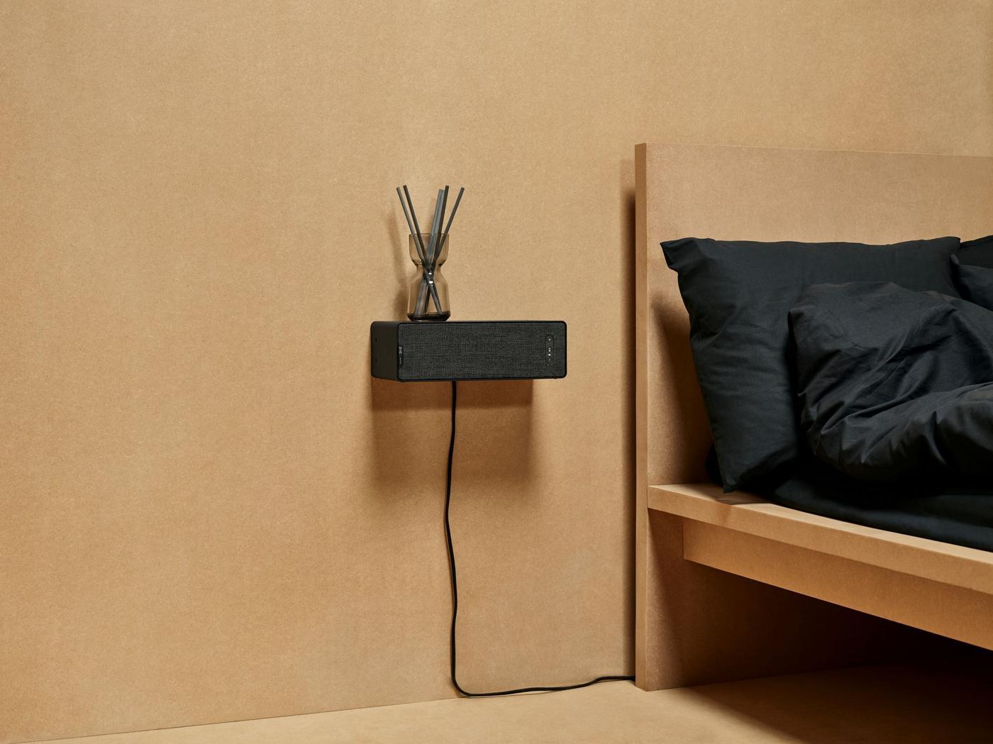 IKEA SYMFONISK WiFi bookshelf speaker mounted on a wall beside a bed with dark bed linen.