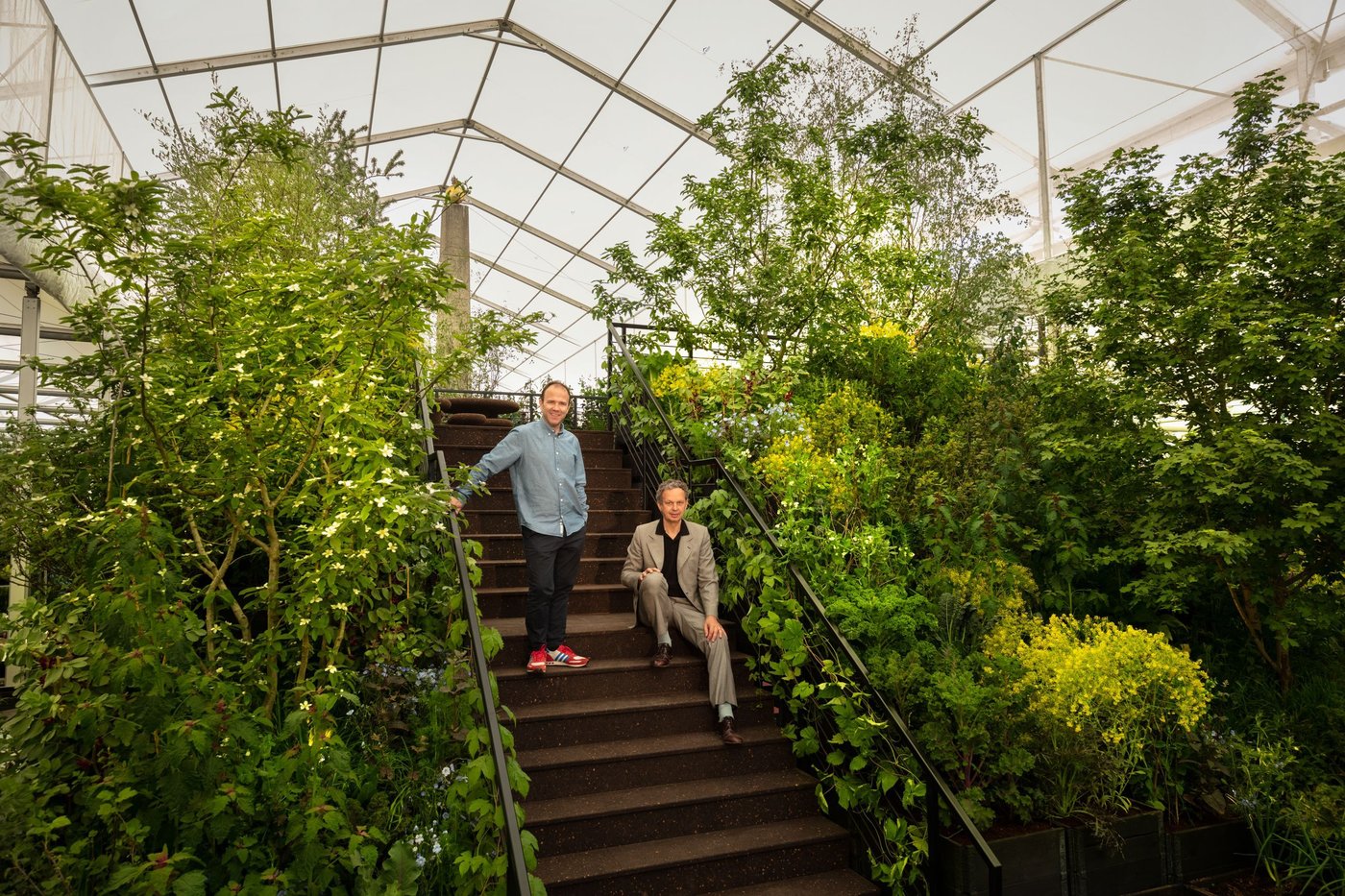 A man sits on the stairs, while the other stands. They are surrounded by plants.