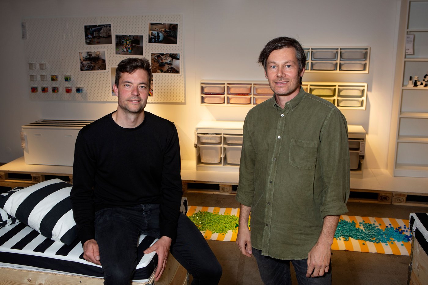 Two men look at the camera. One is standing, while the other perches on the side of a black and white bench.